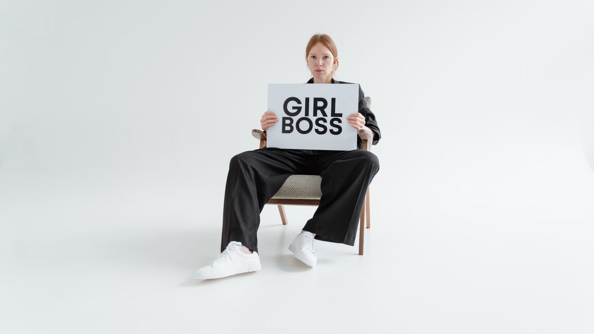 A woman in a black suit holds a 'Girl Boss' sign, exuding confidence on a plain background.
